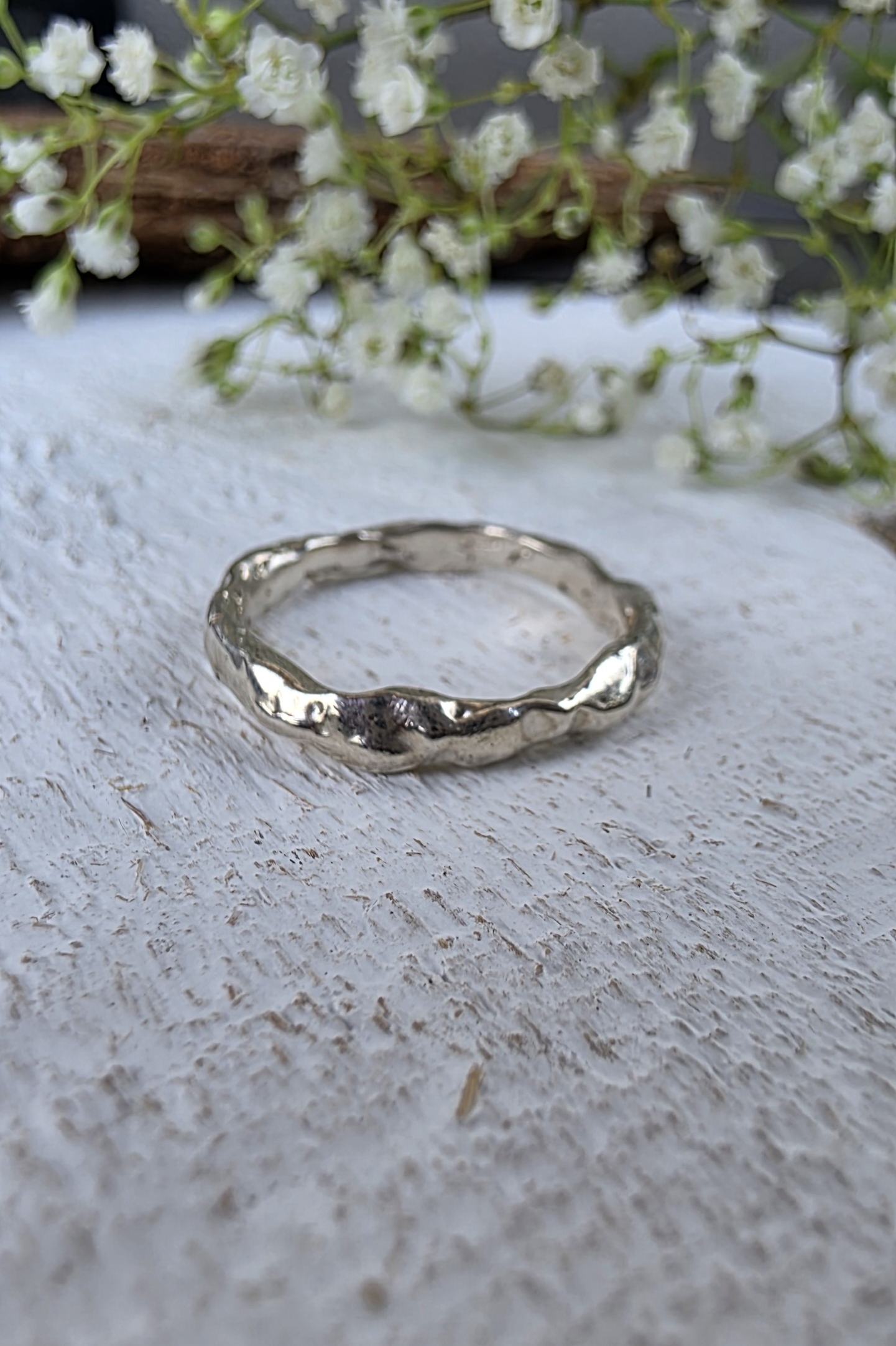 Prospettiva dell'anello Origine in argento lucido su superficie bianca con fiori.