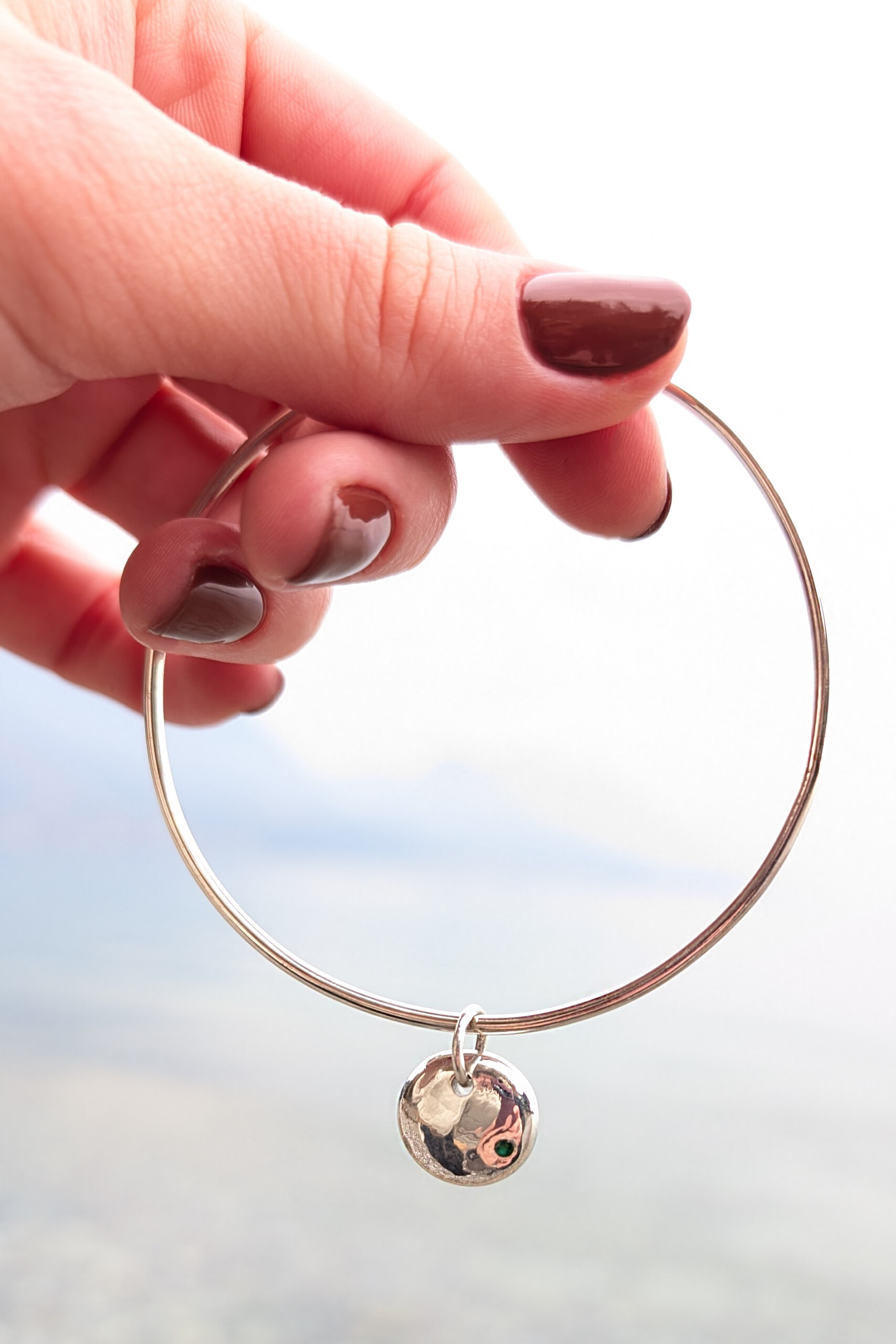 Foto lifestyle della mano di una donna con maglione bianco che indossa il bracciale rigido Pepita con pendente lucido o martellato e pietra naturale verde, con sfondo sfocato di un lago.