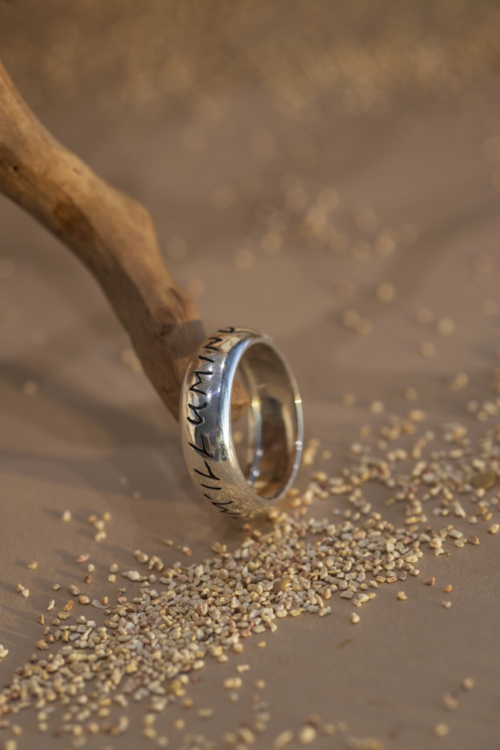 Anello in argento 925 con incisione "M'illumino d'immenso" appoggiato di profilo su legno; un gioiello artigianale che unisce design personalizzato e oreficeria moderna.