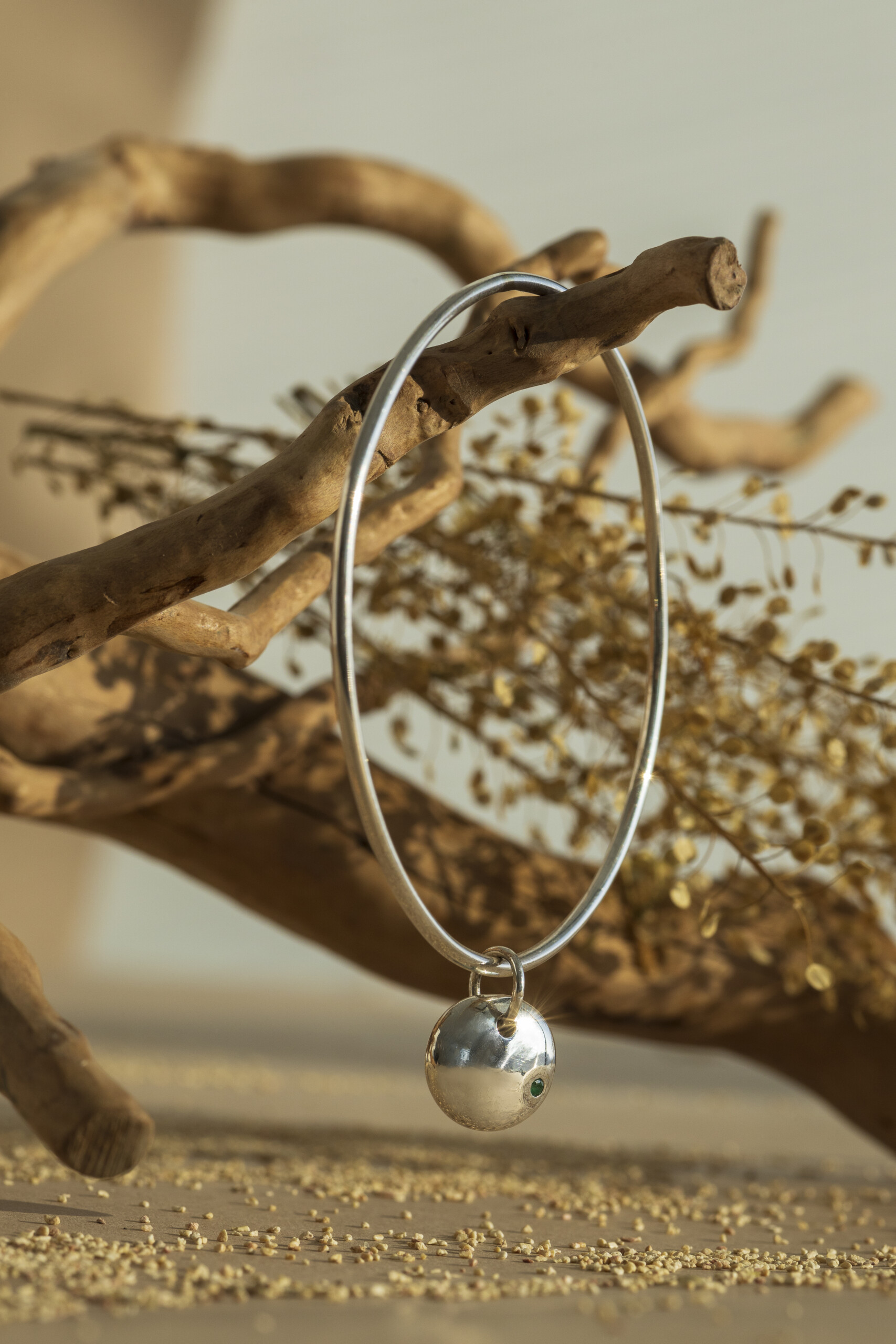 Bracciale rigido artigianale della collezione Pepita in argento 925 lucido, con pendente sferico e smeraldo naturale incastonato o pietra naturale, sospeso su un ramo d'albero.
