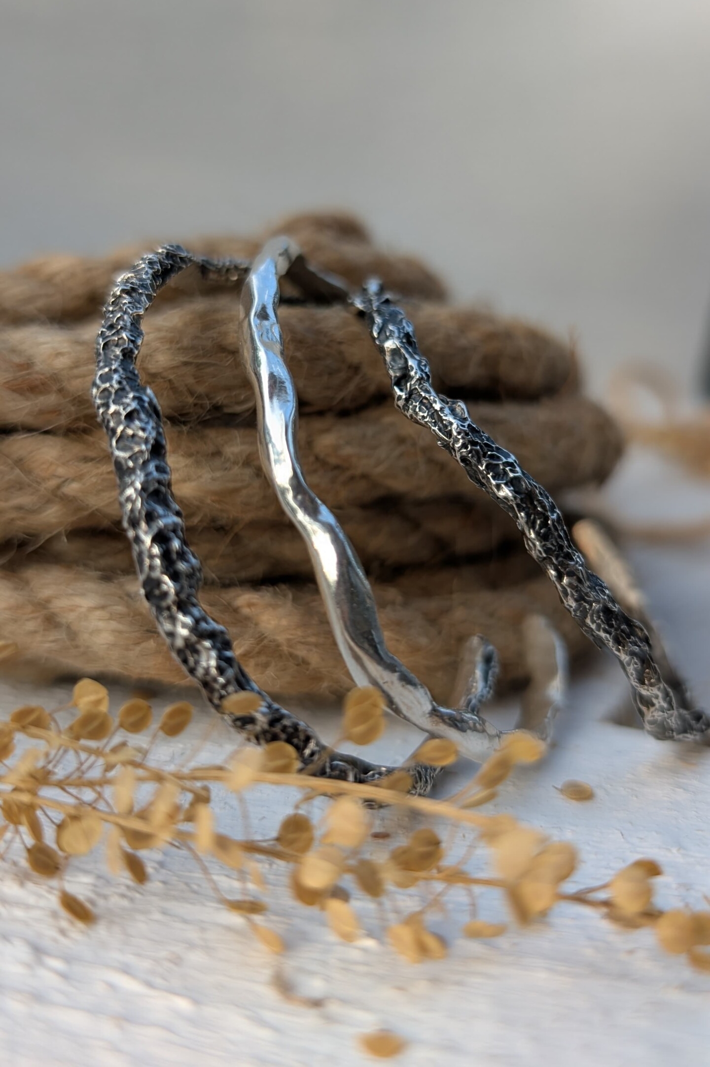 Tre bracciali rigidi in argento artigianali con diverse texture materiche e vulcaniche appoggiati su una corda naturale.