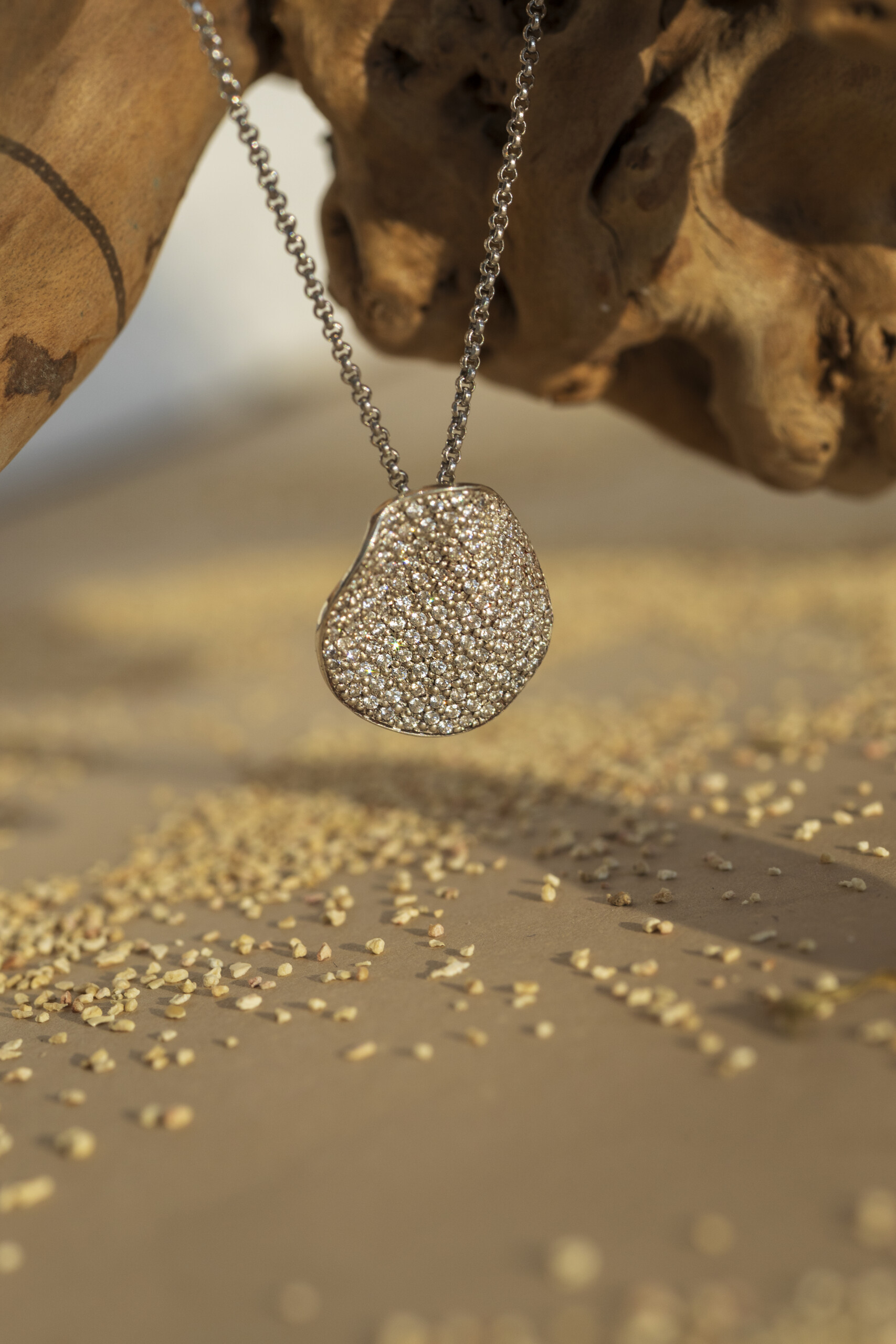 Pendente circolare irregolare con pavé fitto di pietre luminose della collezione Moon, con catena in argento, ispirato alla luna, su sfondo naturale caldo