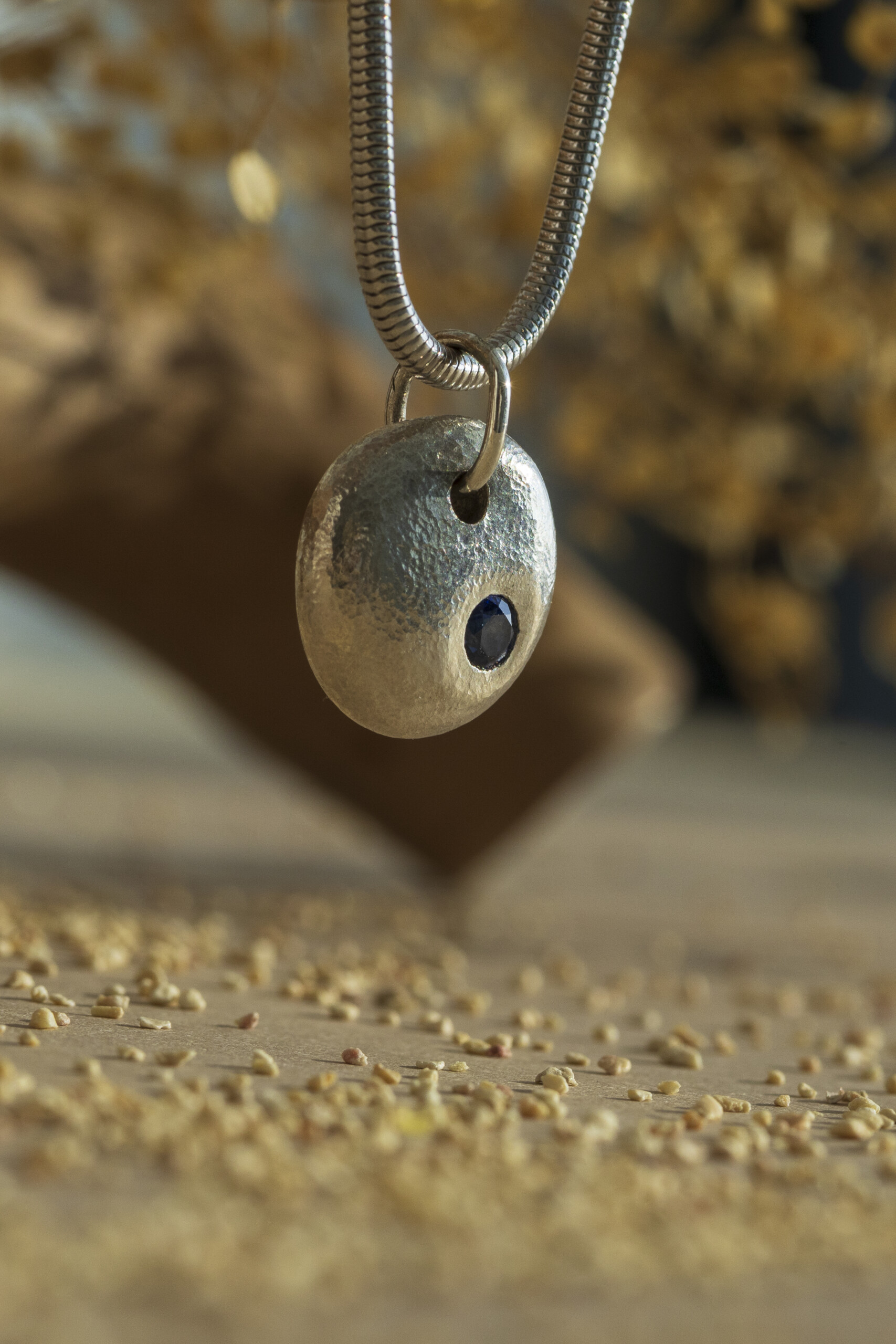 Dettaglio ravvicinato di un ciondolo a forma di pepita in argento martellato con una piccola pietra naturale zaffiro incastonata.