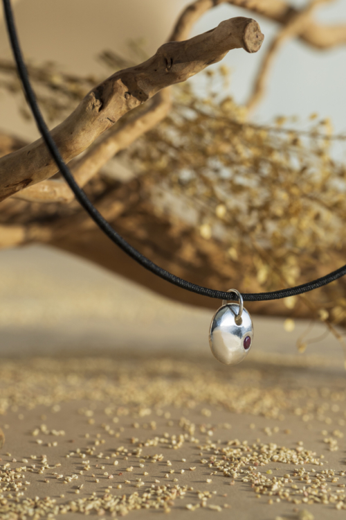 Ciondolo artigianale della collezione Pepita in argento lucido con rubino rosso, montato su un cordino rigido in tessuto nero, sospeso in un set naturale.