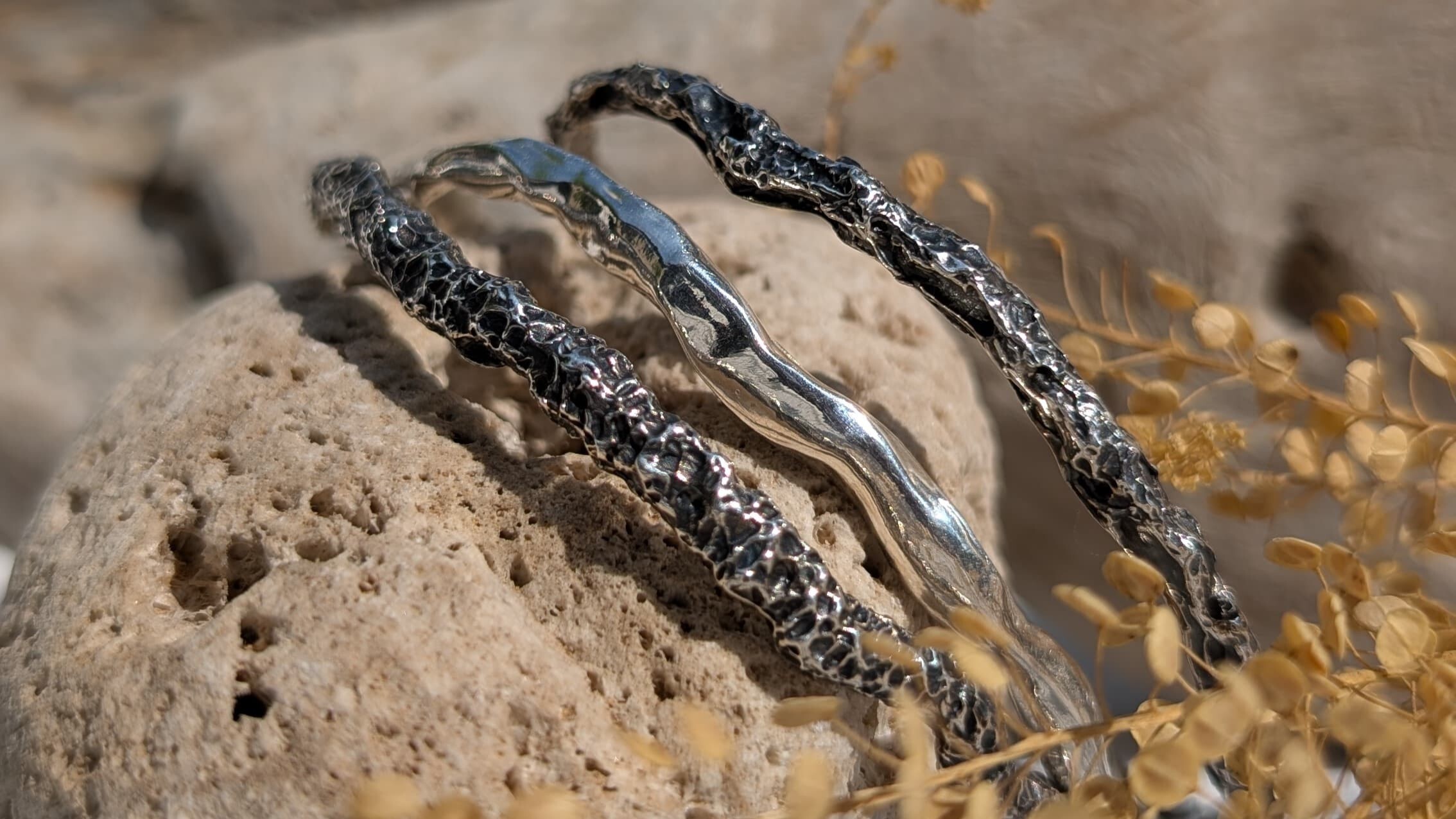 Tre bracciali rigidi artigianali in argento con diverse texture, da lucida a vulcanica scura, esposti su una roccia chiara con elementi naturali.