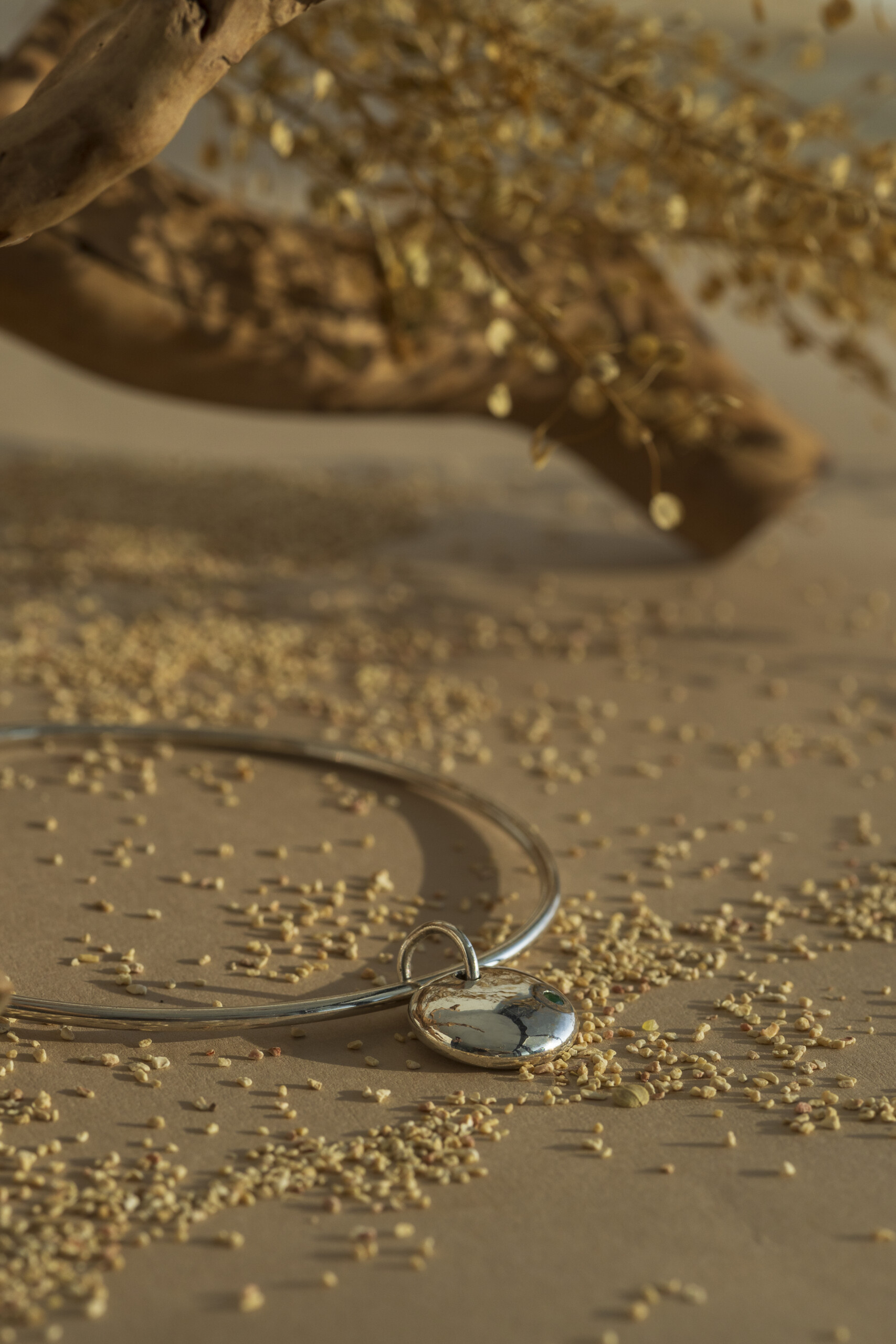 Primo piano del pendente lucido a forma di pepita con piccola pietra naturale verde, appoggiato su una superficie di sabbia chiara con elementi botanici sfocati.