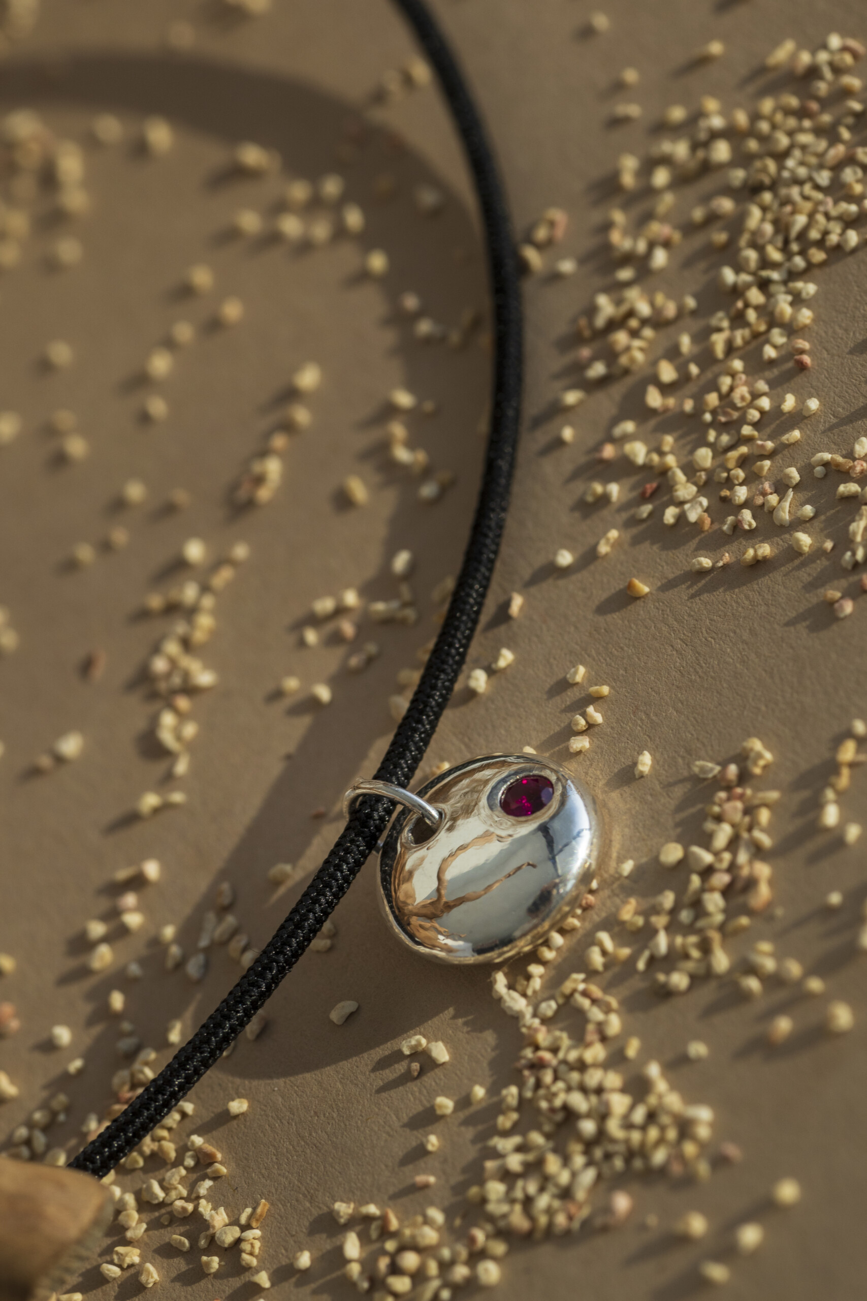 Macro del ciondolo Pepita in argento che mostra la lucentezza della superficie e il taglio del rubino rosso, circondato da piccoli granelli di sabbia.