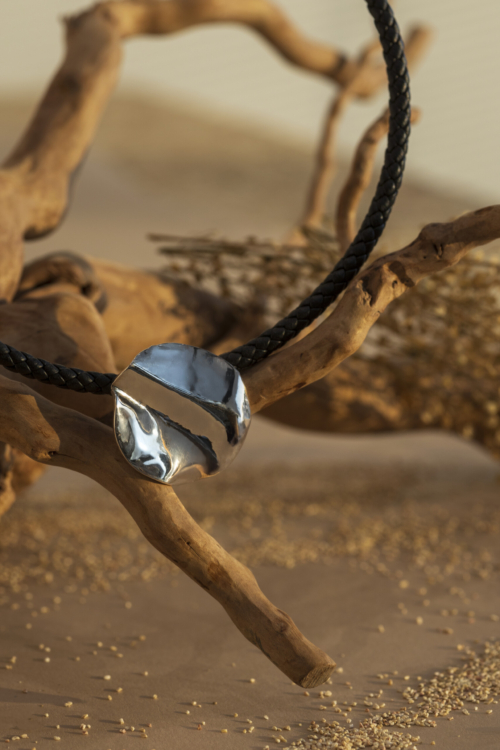 Pendente a lastra ondulata in argento lucido della collezione Moon, montato su un girocollo in pelle nera intrecciata e sospeso su un ramo naturale.