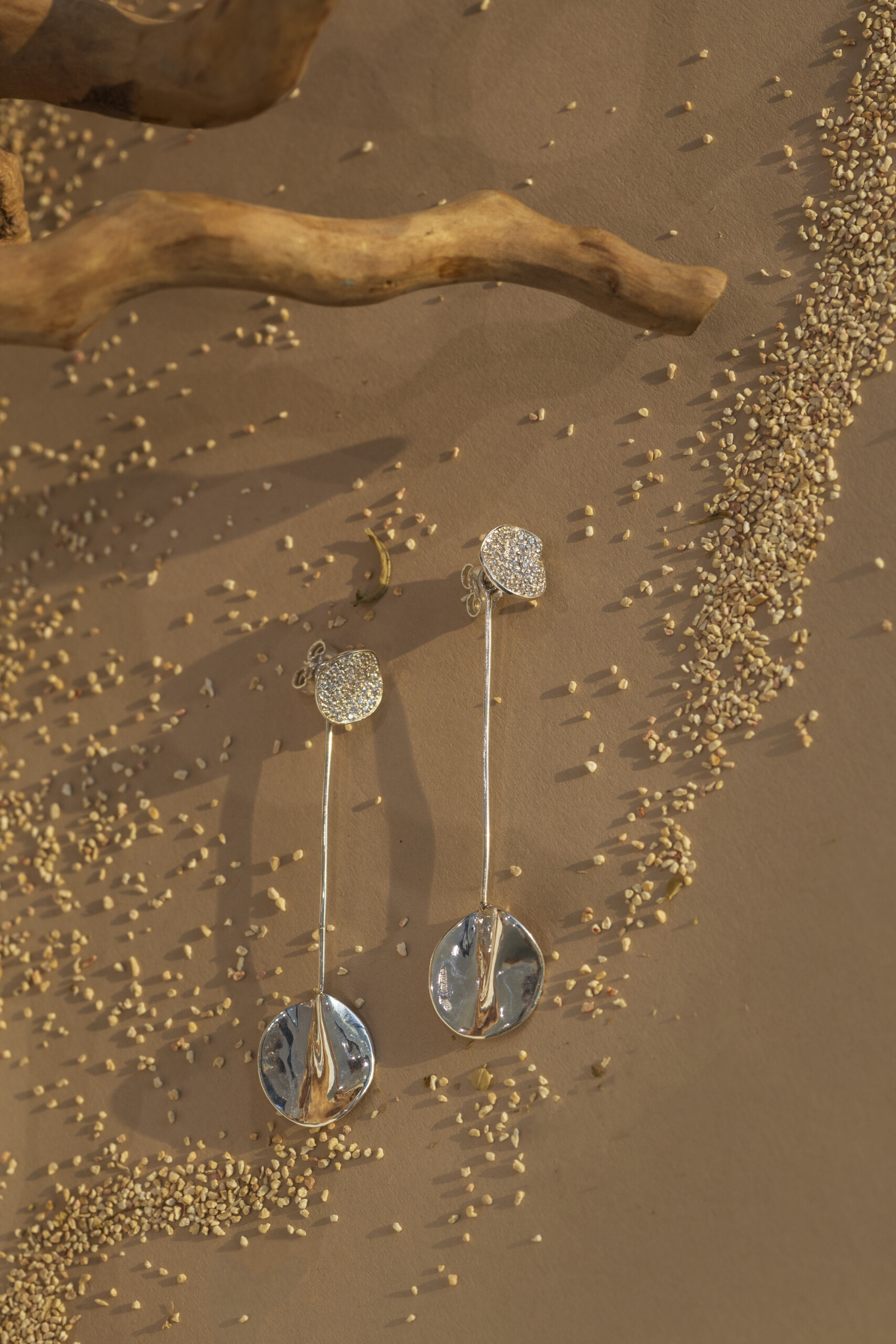 Vista dall'alto degli orecchini Moon pendenti su fondo sabbioso; un esempio di gioielleria artigianale che unisce forme geometriche e texture organiche modellate a mano.