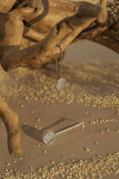 Coppia di orecchini a monachella squadrata della collezione Moon; un pendente è appeso a un ramo e l'altro è poggiato su sabbia, mostrando il pavé di zirconi.